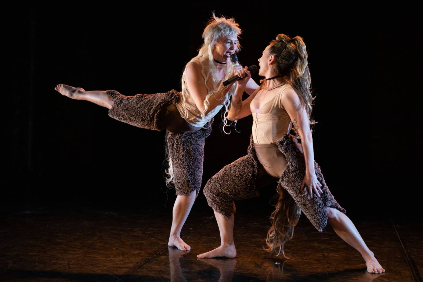 Two women, one lunging, the other in arabesque with mullets talk into microphones